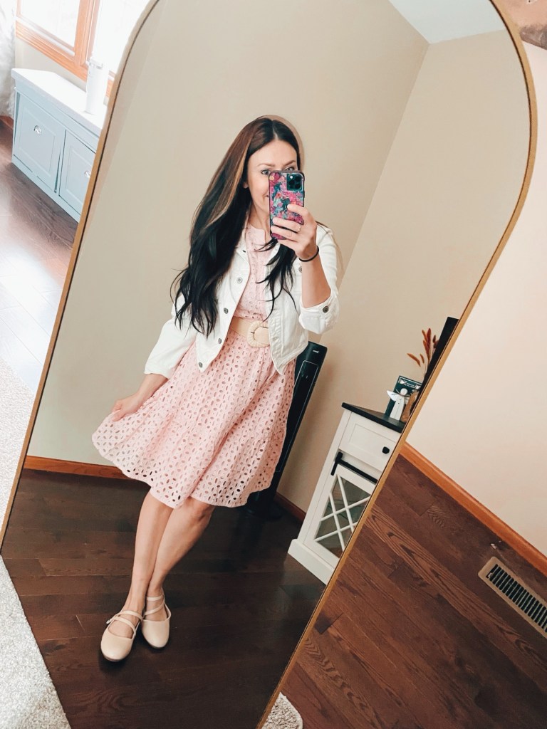 Woman with brown hair wearing a light pink dress, white denim jacket and beige Mary Jane flats with straps taking a photo in front of an arch mirror.